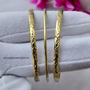 Puede incluir: Tres brazaletes dorados dispuestos verticalmente. Los dos brazaletes exteriores presentan un intrincado patrón de enredadera floral en relieve, mientras que el brazalete central es liso y sencillo. El texto SILVERLEGANCEJAIPUR aparece a la izquierda.