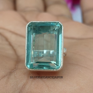May include: A silver ring featuring a large, rectangular, faceted light blue gemstone set in a smooth metal bezel. The stone is transparent with visible internal inclusions. The text SILVERLEGANCEJAIPUR is printed below the ring setting.