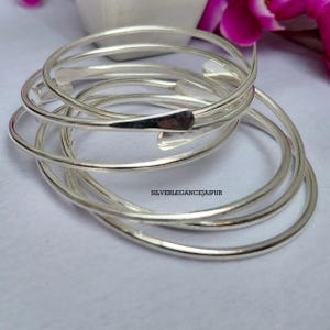 May include: A set of stacked silver-colored spiral bangles featuring a smooth, polished finish and tapered ends. The text SILVERLEGANCEJAIPUR is printed in the center of the coiled metal bands, which are arranged in a circular, overlapping formation.