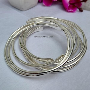 May include: A set of multiple interlocking silver-colored bangles with a smooth, polished finish. The bracelets are arranged in a circular, overlapping formation. The text SILVERELEGANCEJAIPUR is printed in black in the center of the bangles.