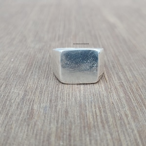Peut inclure: Une bague chevalière en argent dotée d'une face rectangulaire plate avec une finition texturée et mate. L'anneau est lisse et poli, et le texte SILVERLEGANCEJAIPUR est visible en petites lettres sombres près du bord supérieur de la bague.