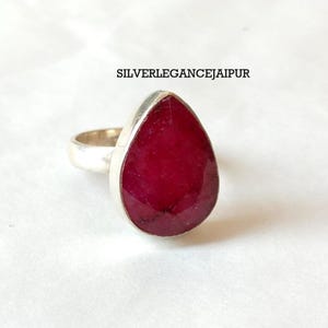 Puede incluir: Un anillo de plata con una gran piedra preciosa de color rojo intenso, tallada en forma de pera y engastada en un bisel de metal liso. El texto SILVERLEGANCEJAIPUR es visible sobre el anillo. La banda es sencilla y metálica, con un diseño básico.