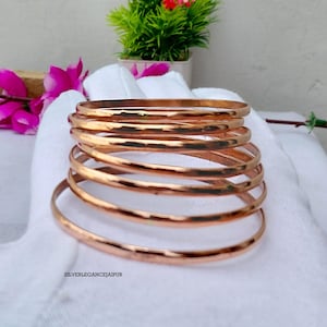 May include: A stack of six polished copper bangles with a smooth, rounded surface. The bracelets are arranged in a vertical column, showcasing a warm, metallic reddish-brown tone. The text "SILVERELEGANCEJAIPUR" is printed in small black letters.