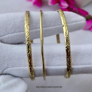 Pode incluir: Três pulseiras douradas, sendo que as duas pulseiras externas apresentam um padrão de videira floral em relevo e a pulseira central tem uma superfície lisa e simples. O texto SILVERELEGANCEJAIPUR está impresso em preto na imagem.