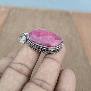 May include: An oval-shaped pendant featuring a faceted, deep pink gemstone set in a decorative sterling silver frame with intricate filigree detailing along the sides. The silver bail is stamped with 925, indicating sterling silver composition.