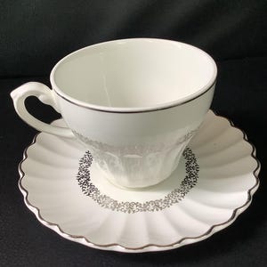 May include: A white porcelain teacup and saucer set featuring a delicate silver filigree pattern along the rim of the cup and a matching intricate silver floral border on the scalloped saucer, finished with a thin silver trim on all edges.