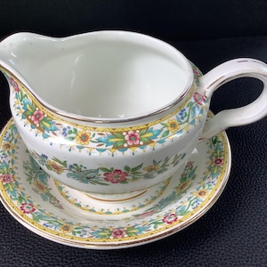 Peut inclure: Une saucière en céramique blanche avec sa soucoupe assortie, ornée d'un motif floral coloré dans des tons de rose, jaune, bleu et vert, rehaussée d'un liseré doré sur les bords de l'anse, du bec verseur et de la base circulaire du récipient.