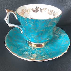 Peut inclure: Un ensemble tasse et soucoupe bleu sarcelle orné d'un motif floral et végétal en or métallique. La tasse présente un bord festonné, une anse dorée et un intérieur blanc avec des détails dorés sur le bord. La soucoupe porte le même motif.