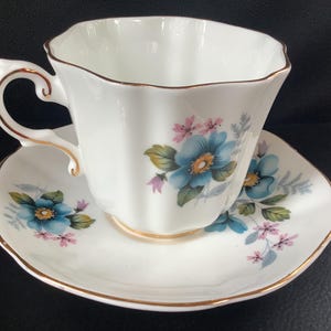 Peut inclure: Un ensemble tasse et soucoupe en porcelaine blanche avec un bord festonné et un liseré doré. L'ensemble est orné d'un motif floral composé de fleurs bleues, de feuilles vertes et de petites fleurs roses. La tasse possède une anse incurvée dorée.