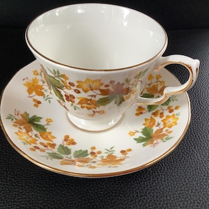 May include: A white porcelain teacup and saucer set featuring a delicate pattern of autumn-colored maple leaves, yellow flowers, and small berries. Both the cup and the saucer are finished with a thin gold trim along the scalloped edges.