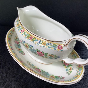 Peut inclure: Une saucière en céramique blanche avec soucoupe ovale intégrée, ornée d'un motif floral coloré de fleurs rouges, jaunes et bleues avec des feuilles vertes, finie par un fin liseré doré sur le bord et l'anse incurvée.