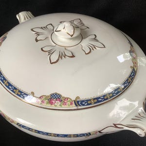 Peut inclure: Une soupière ronde en céramique couleur crème avec son couvercle assorti. Le couvercle présente un bouton central orné de motifs de feuilles dorées. Le bord est décoré d'une bordure bleue et dorée, agrémentée de petites fleurs roses.