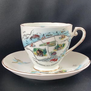 Peut inclure: Une tasse à thé et sa soucoupe en porcelaine blanche ornées d'une carte illustrée de la région de la Gaspésie au Québec, Canada. Le motif comprend des dessins d'ours, de cerf, de phares et des étiquettes textuelles. Bordure dorée.