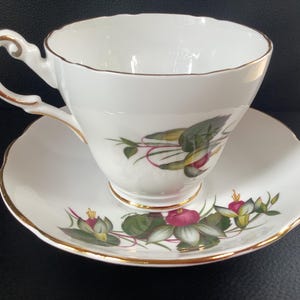 Peut inclure: Un ensemble tasse et soucoupe en porcelaine blanche orné d'un motif floral délicat avec des feuilles vertes et des boutons roses. Le bord de la tasse, l'anse et le bord de la soucoupe sont rehaussés d'un fin liseré doré, avec des bords festonnés.