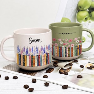 May include: Two ceramic mugs with speckled finishes. The left mug is off-white with the name Susan and a row of colorful books topped with purple flowers. The right mug is sage green with the name mina and a row of books topped with pink flowers.