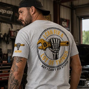 May include: A light gray short-sleeve t-shirt featuring a graphic on the back of a skeleton hand holding a yellow wrench inside a circle with the text I CAN FIX A LOT OF THINGS BUT I CAN'T FIX STUPID. A smaller matching graphic is on the sleeve.