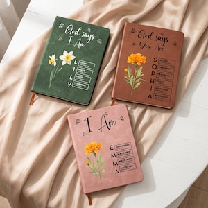 May include: Three rectangular journals in dark green, brown, and dusty pink. The green journal features white daffodils and the text God says I Am LILY. The brown journal shows an orange flower and the text God says You Are SOPHIA. The pink journal displays an orange flower and the text I Am EMMA with various scripture references.