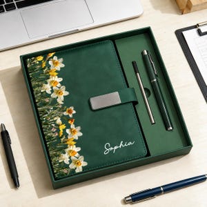 Peut inclure: Un ensemble carnet vert foncé avec un imprimé floral de jonquilles blanches et jaunes sur le côté gauche, un fermoir en métal argenté et le prénom Sophia écrit en blanc. L'ensemble comprend un stylo vert foncé assorti et un stylet argenté fin.