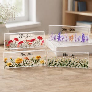 Peut inclure: Quatre blocs rectangulaires en acrylique transparent contenant des fleurs pressées et des noms personnalisés. Un bloc affiche des œillets rouges avec Mrs. Jessica, un des fleurs violettes avec Mrs. Anna, un des soucis jaunes avec Mrs. Alice et un des fleurs blanches avec Mrs. Lily.
