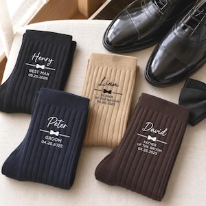 May include: Four pairs of ribbed socks in black, navy, tan, and dark brown. Each pair features a white printed name, a small bow tie icon, a role such as Best Man, Groom, or Father of the Groom, and a specific wedding date. Black dress shoes.
