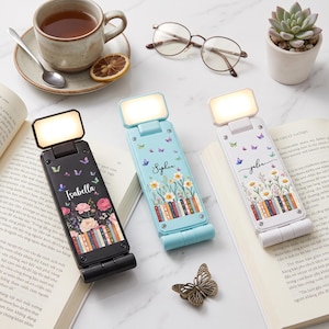May include: Three rectangular foldable LED reading lights with illuminated panels. Each light features a unique floral and book-themed graphic with the names Isabella, Sophia, and Julia printed in script font on black, light blue, and white bases.