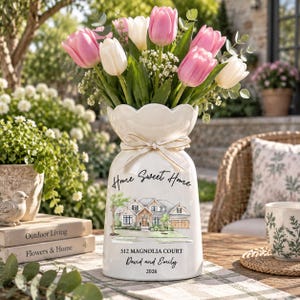 May include: A white ceramic vase with a scalloped rim and a cream ribbon bow, filled with pink and white tulips. The vase features a printed illustration of a house, the text Home Sweet Home, 512 MAGNOLIA COURT, David and Emily, and 2026.