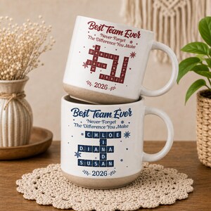 May include: Two ceramic mugs stacked vertically, featuring a beige base and white upper. The top mug has red text reading Best Team Ever, Never Forget The Difference You Make, and 2026 with a crossword design. The bottom mug has blue text and design.