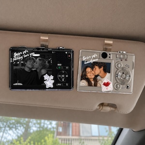 May include: Two rectangular car visor clips shaped like digital cameras. The black camera features a black-and-white photo of a couple and the text Drive safe, I fucking love you. The silver camera shows a photo of a couple and the text Drive safe, handsome.
