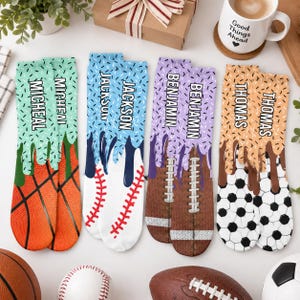 May include: Four pairs of crew socks featuring sports themes with personalized names. The socks display basketball, baseball, football, and soccer ball patterns with a dripping icing effect in green, blue, purple, and tan, labeled Micheal, Jackson, Benjamin, and Thomas.