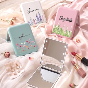May include: Three rectangular compact mirrors with floral designs and names Jasmine, Elizabeth, and Sophia printed on the covers. One open mirror features integrated LED lights around the frame. Two lipsticks and a pair of pearl earrings are nearby.