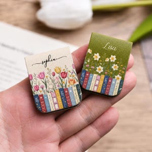 May include: Two rectangular magnetic bookmarks with a textured finish. One is cream-coloured with a floral print of tulips and the name Sophia, the other is olive green with a print of white daffodils and the name Lisa. Both feature book spines.