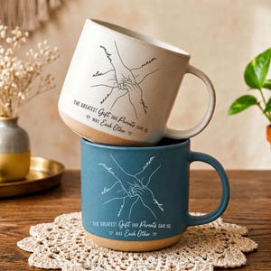 May include: A cream-coloured ceramic mug stacked on a blue ceramic mug. Both feature a line art illustration of hands joined together with the names Jackson, Mason, Sophia, Anna, and Alex, and the text: The greatest gift our parents gave us was each other.