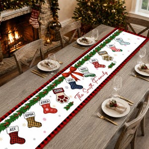 May include: A white table runner with a red and black buffalo plaid border, featuring a green garland with a red bow and multiple illustrated Christmas stockings. Each stocking is personalized with a name, including The Smith Family text.