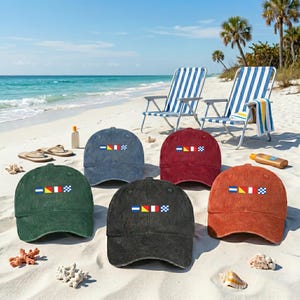 May include: Six corduroy baseball caps in various colors: green, blue, red, black, and orange. Each cap features a nautical flag design embroidered on the front. The caps are displayed on a sandy beach with beach chairs and ocean in the background.