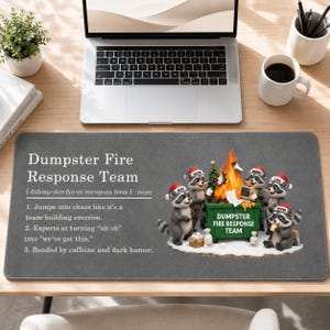 May include: A grey desk mat featuring the text Dumpster Fire Response Team with a definition and a graphic of five raccoons wearing Santa hats gathered around a green dumpster with a fire inside, labelled Dumpster Fire Response Team.