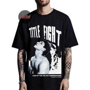Puede incluir: Camiseta negra con un gr&aacute;fico que presenta el texto "TITLE FIGHT" en letras blancas en negrita. Debajo del texto hay una imagen en blanco y negro de una persona mirando hacia arriba. El texto "LIVE AT THE VELVET UNDERGROUND 506 QUEEN ST W TORONTO" est&aacute; en la parte inferior.