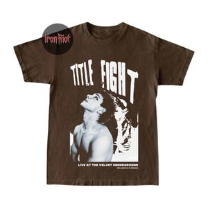 Puede incluir: Camiseta marr&oacute;n con un gr&aacute;fico en blanco y negro de una persona mirando hacia arriba, con las palabras "TITLE FIGHT" en letras blancas grandes. El texto "LIVE AT THE VELVET UNDERGROUND" est&aacute; debajo.