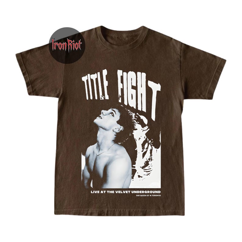 Puede incluir: Camiseta marr&oacute;n con un gr&aacute;fico en blanco y negro de una persona mirando hacia arriba, con las palabras "TITLE FIGHT" en letras blancas grandes. El texto "LIVE AT THE VELVET UNDERGROUND" est&aacute; debajo.