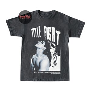 Puede incluir: Camiseta gris oscuro con un gr&aacute;fico en blanco y negro de una persona mirando hacia arriba, con las palabras "TITLE FIGHT" en letras blancas grandes. Debajo del gr&aacute;fico, el texto dice "LIVE AT THE VELVET UNDERGROUND". La camiseta tiene un aspecto vintage.