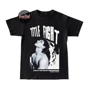 Puede incluir: Camiseta negra con un dise&ntilde;o gr&aacute;fico que presenta el texto "TITLE FIGHT" en letras blancas grandes. El dise&ntilde;o incluye una imagen en blanco y negro de una persona mirando hacia arriba. El texto "LIVE AT THE VELVET UNDERGROUND" tambi&eacute;n est&aacute; impreso en la camiseta.