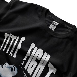 Puede incluir: Camiseta negra con las palabras "TITLE FIGHT" en letras blancas grandes. Una imagen en escala de grises de la cara de una persona est&aacute; debajo del texto. La camiseta tiene cuello redondo y una etiqueta "GILDAN".