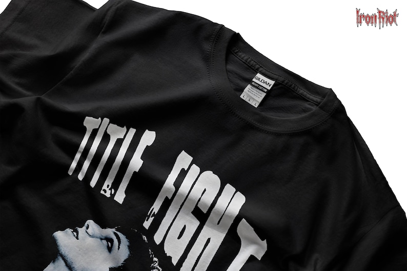 Puede incluir: Camiseta negra con las palabras "TITLE FIGHT" en letras blancas grandes. Una imagen en escala de grises de la cara de una persona est&aacute; debajo del texto. La camiseta tiene cuello redondo y una etiqueta "GILDAN".