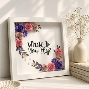 May include: A square white shadow box frame containing the black text What If You Fly? surrounded by paper flowers in shades of purple, pink, and beige, accented with small green leaves and white pearl beads arranged in a corner floral design.