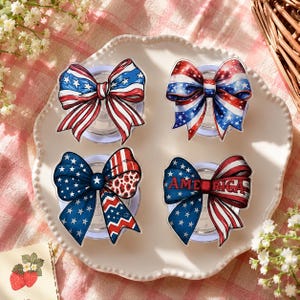May include: Four decorative bow-shaped suction cup designs in red, white, and blue, featuring stars and stripes patterns. One bow has the word "AMERICA" on it. The bows are arranged on a white, scalloped plate, evoking a patriotic theme.