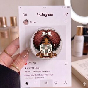 May include: A circular phone grip featuring an illustration of a person with voluminous dark curly hair and a white bow, holding an open book with the text 'JUST A GIRL WHO LOVES BOOKS' printed on the cover, displayed on a printed Instagram interface.