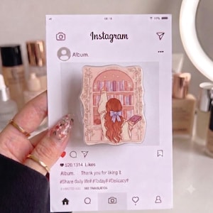 May include: A phone grip featuring an illustration of a person with long wavy hair and a purple bow reaching for a book on a shelf. The grip is mounted on a white card designed to look like an Instagram post with the text Album, 520,1314 Likes, and hashtags.