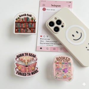 May include: Several phone grips with book-related designs are shown. One features a bookshelf and the text "book tok." Another says "Born to Read Forced to Work." A third grip says "Booked and Busy." A phone case with a smiley face is also present.