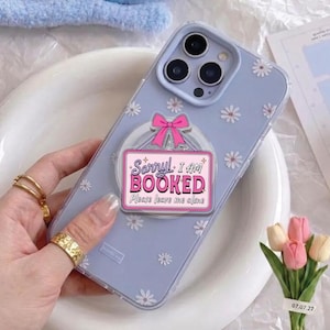 May include: A light purple phone case with a daisy pattern and a pop-up grip. The grip features a pink sign with the text "Sorry! I AM BOOKED Please leave me alone". The phone case is on a white plate.