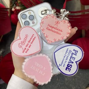 May include: A clear phone case with four heart-shaped pop sockets. Each pop socket has a different message in pink and white, including "Please skip the flowers buy me books."