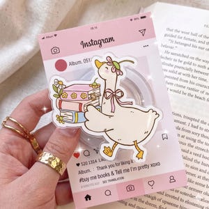 Może przedstawiać: Różowa naklejka w stylu Instagrama z kreskówkową gęsią niosącą książki i noszącą wianek z kwiatów. Naklejka zawiera tekst "#buy me books & Tell me I'm pretty xoxo" i jest trzymana przez dłoń ze złotymi pierścionkami.